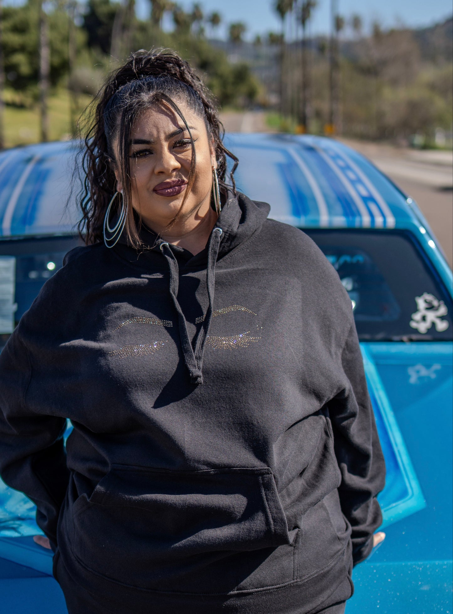 Person wearing a black hoodie and pants standing in front of a blue car with a desert landscape in the background.