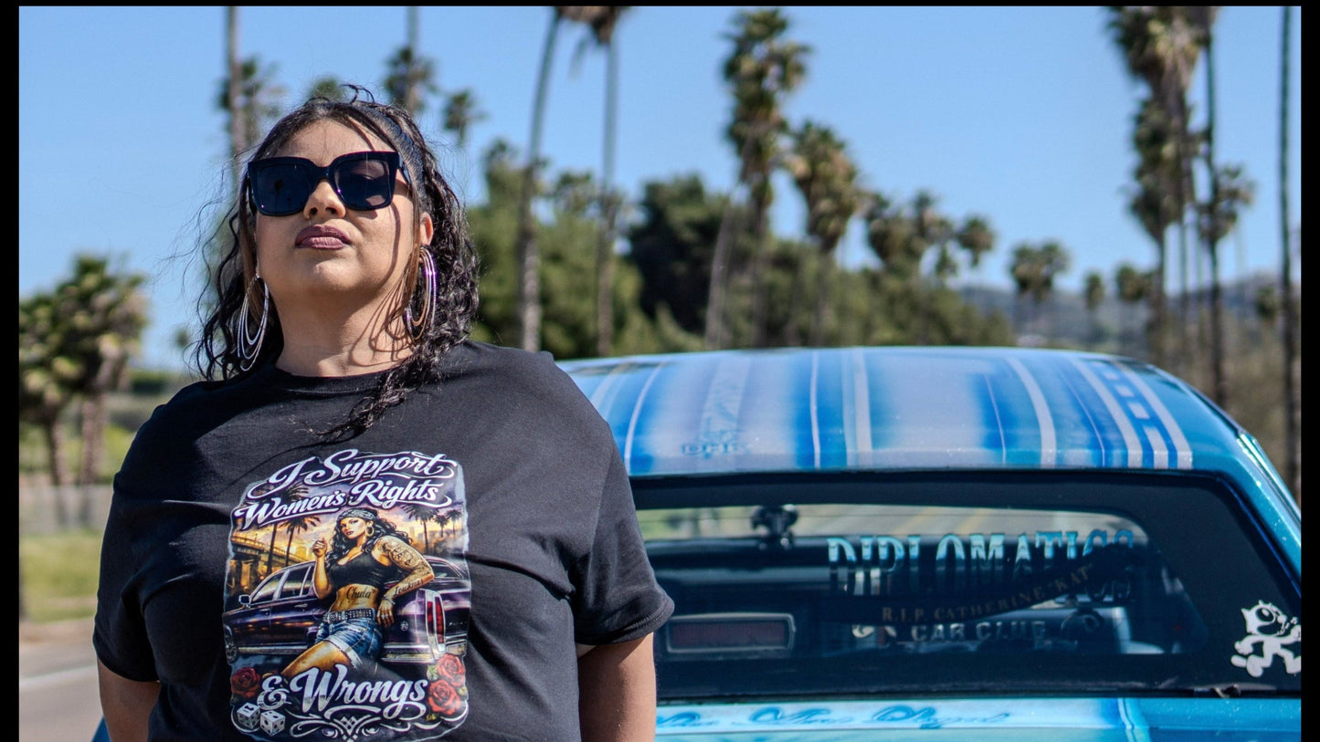 Person wearing sunglasses and a graphic t-shirt standing next to a blue car with palm trees in the background