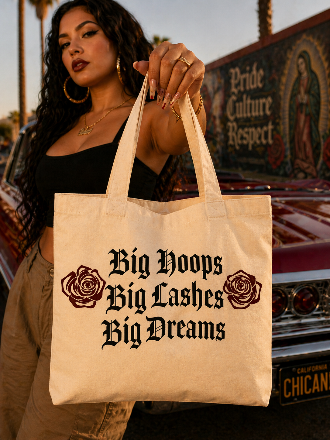 Woman holding a tote bag with 'Big Hoops Big Lashes Big Dreams' text and roses, with a car and mural in the background.