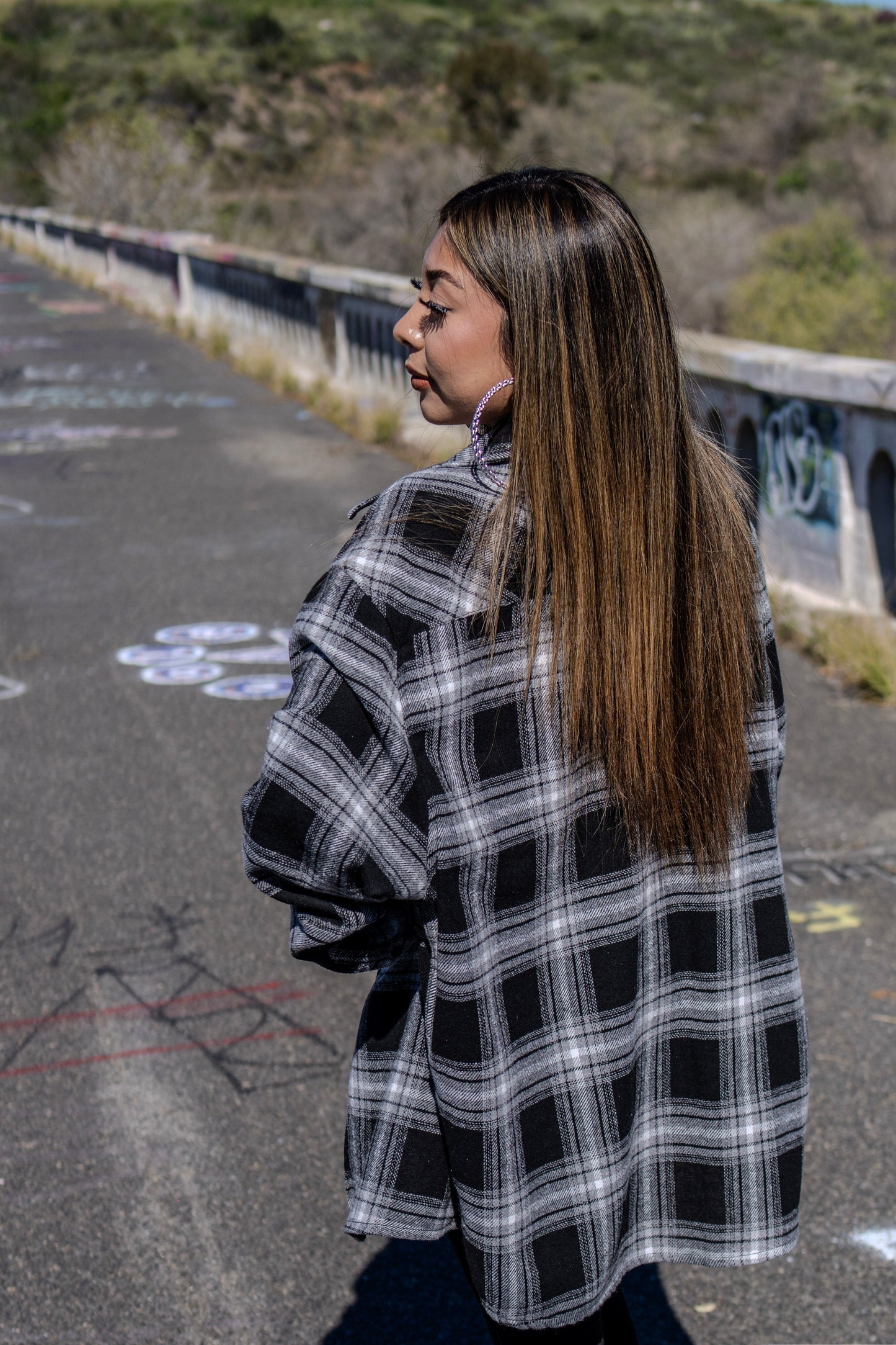 Person wearing a black and white plaid coat standing on a road with a bridge in the background.