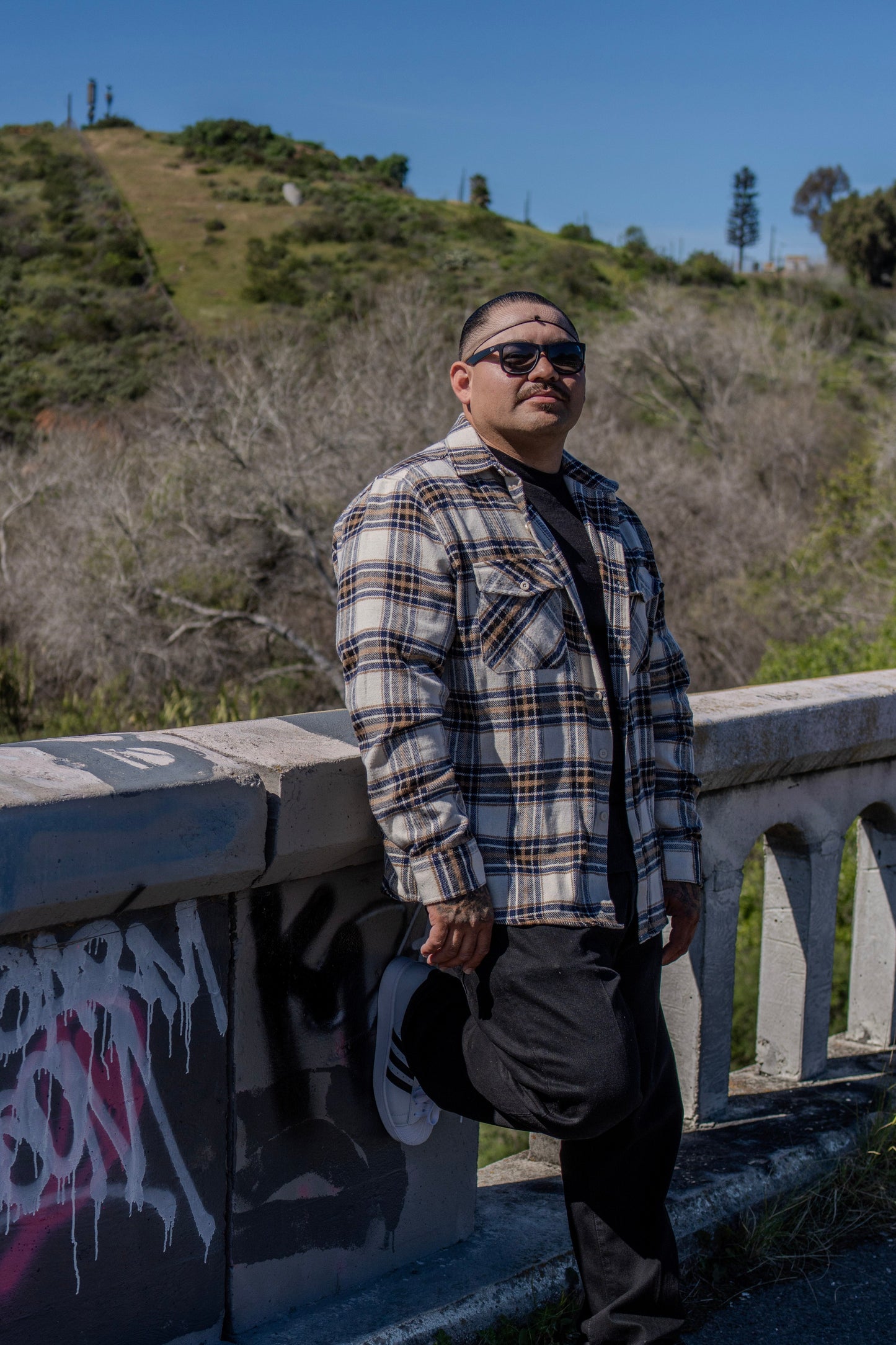 Man wearing sunglasses and a plaid shirt standing on a bridge with graffiti, surrounded by natural scenery.